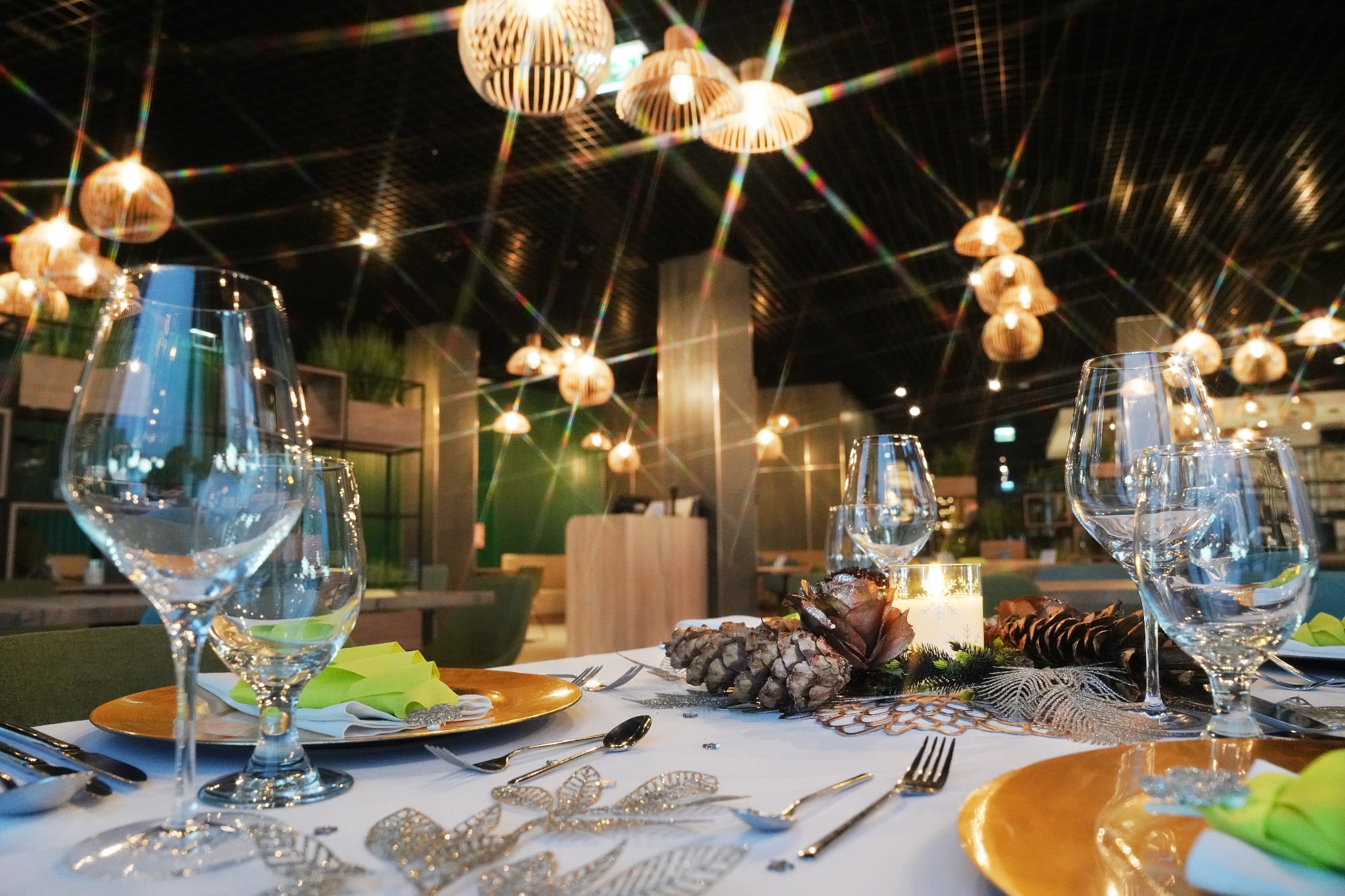 Uroczysta dekoracja stołu z elementami roślinnymi, szyszkami, świecami i elegancką zastawą pod wiszącymi lampami.