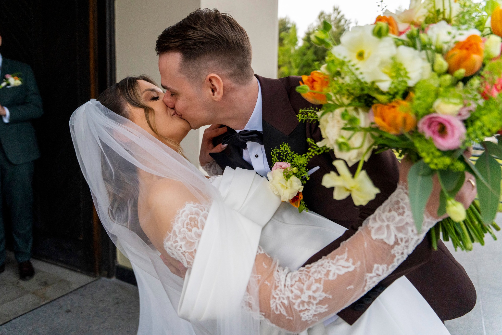 Pocałunek nowożeńców: panna młoda w koronkowej sukni i welonie, trzymana w ramionach przez pana młodego w brązowym smokingu, z bukietem kwiatów na tle jasnego budynku.