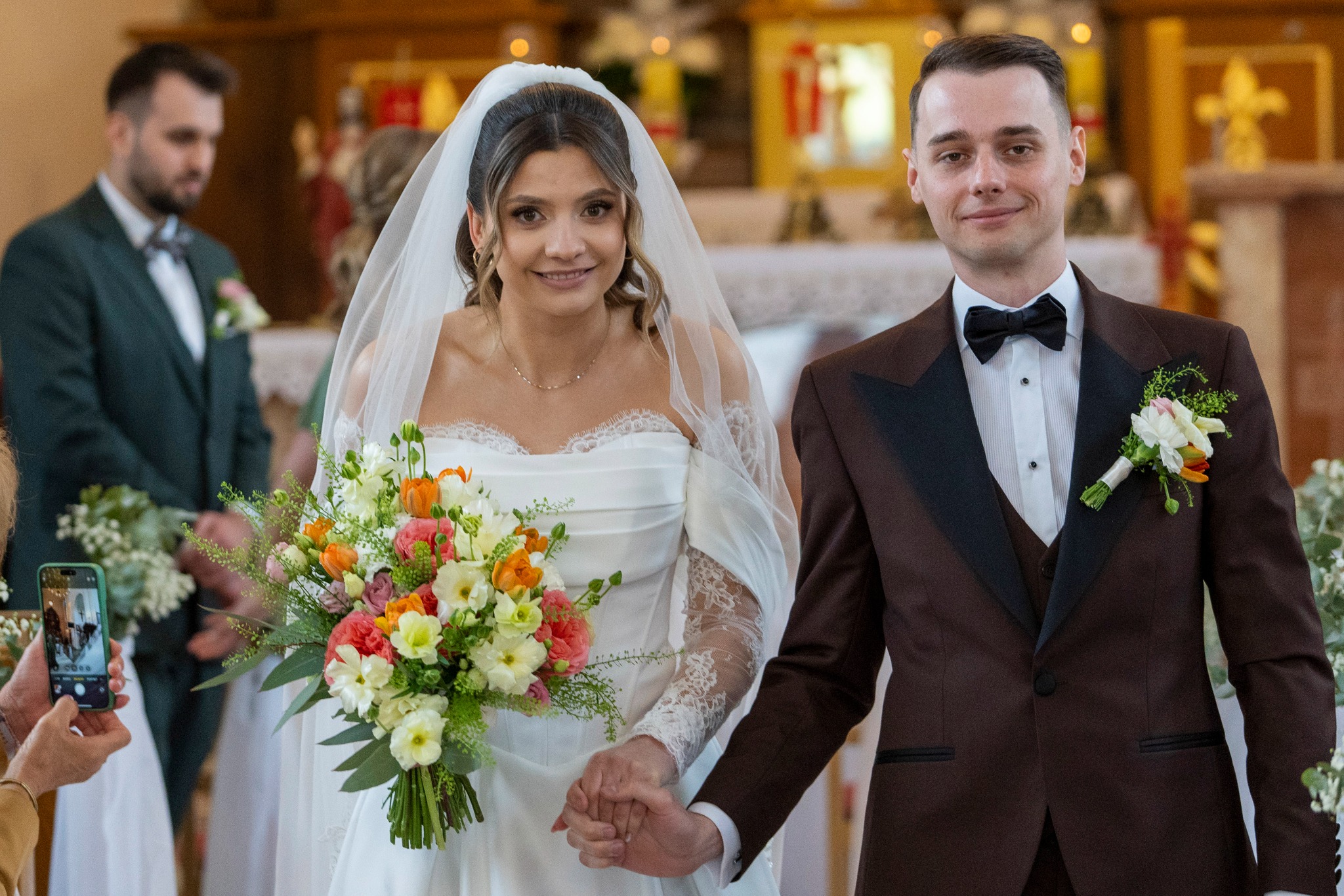 Uśmiechnięta para młoda w kościele, panna młoda z bukietem, pan młody w brązowym smokingu, trzymają się za ręce. W tle goście i ołtarz.