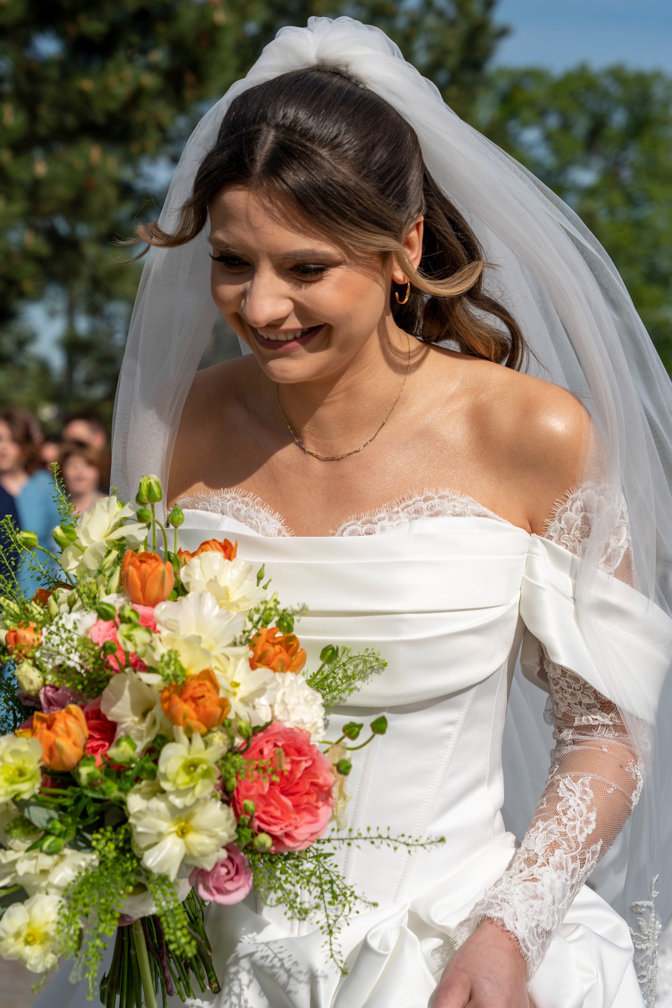 Uśmiechnięta panna młoda w białej sukni z koronkowymi rękawami i welonem, trzymająca bukiet kwiatów w odcieniach bieli, pomarańczy i różu na tle zieleni.