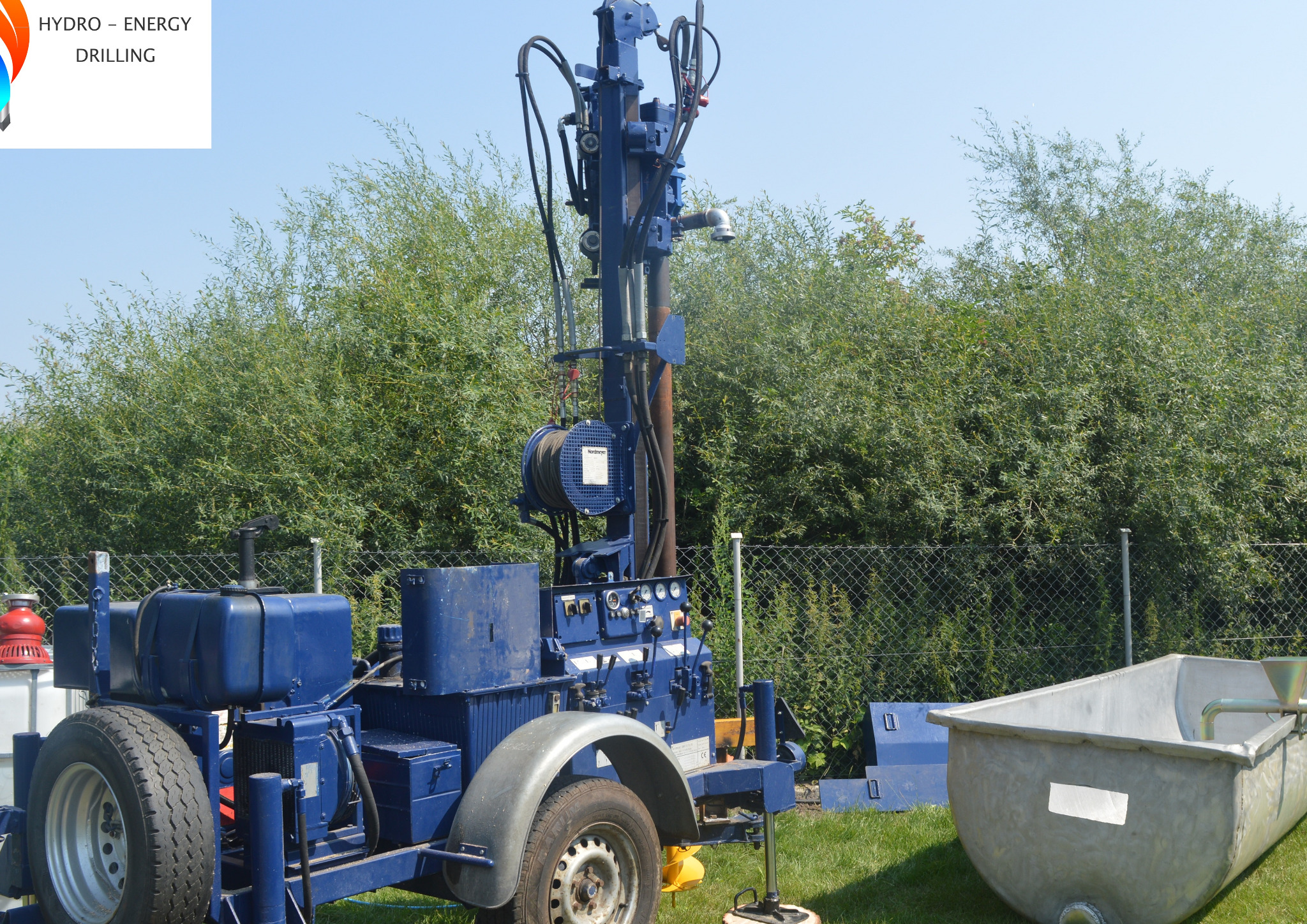Niebieska, mobilna wiertnica na trawie z metalową wanną na płuczkę w tle, widok z boku.
