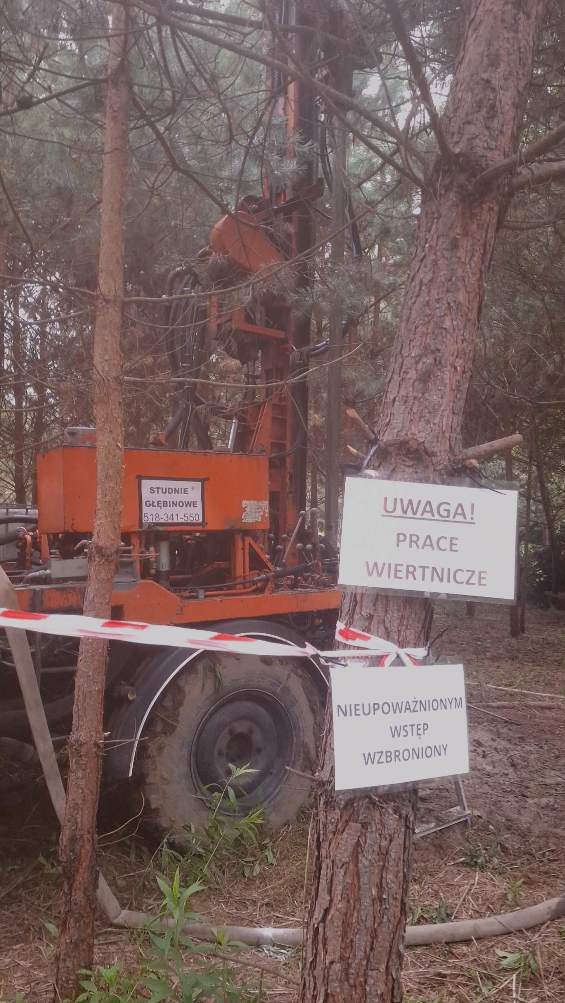 Pomarańczowa wiertnica na tle drzew, z tabliczkami ostrzegawczymi. Widoczne błoto i elementy urządzenia. Ujęcie z poziomu gruntu w lesie.