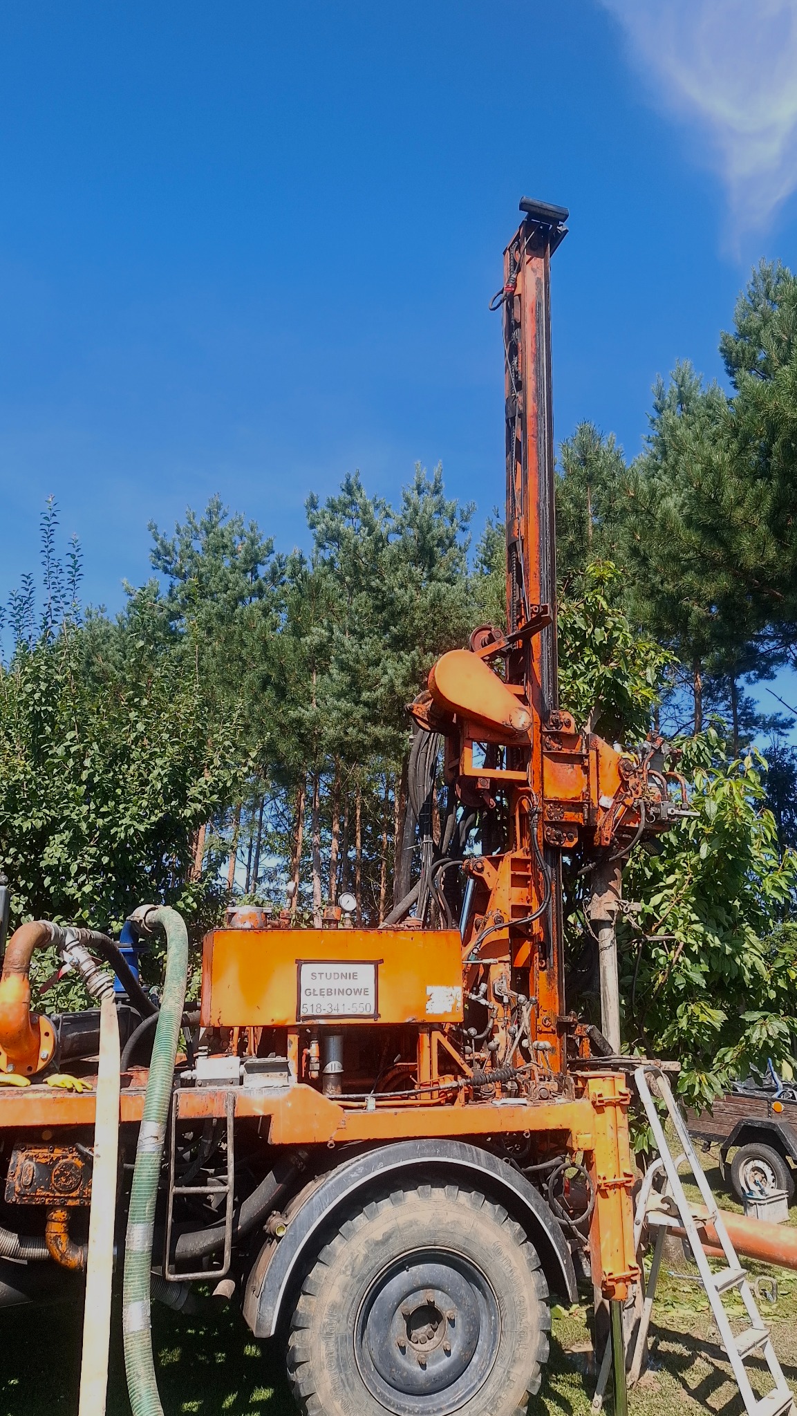Pomarańczowa wiertnica do studni głębinowych na tle zieleni i błękitnego nieba. Widoczne przewody, drabina i opony. Tabliczka z numerem telefonu.