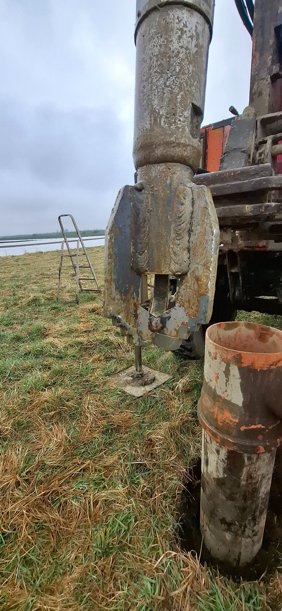 Wiertnica w trakcie pracy na zewnątrz, z widocznym elementem wiercącym i fragmentem drabiny w tle. Sprzęt pobrudzony ziemią.