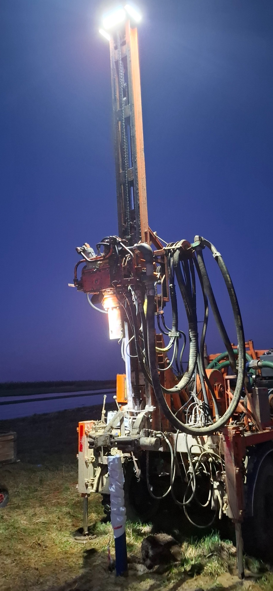Pomarańczowa wiertnica do studni głębinowych oświetlona reflektorem na tle nocnego nieba, widoczna instalacja rur i przewodów, teren trawiasty.