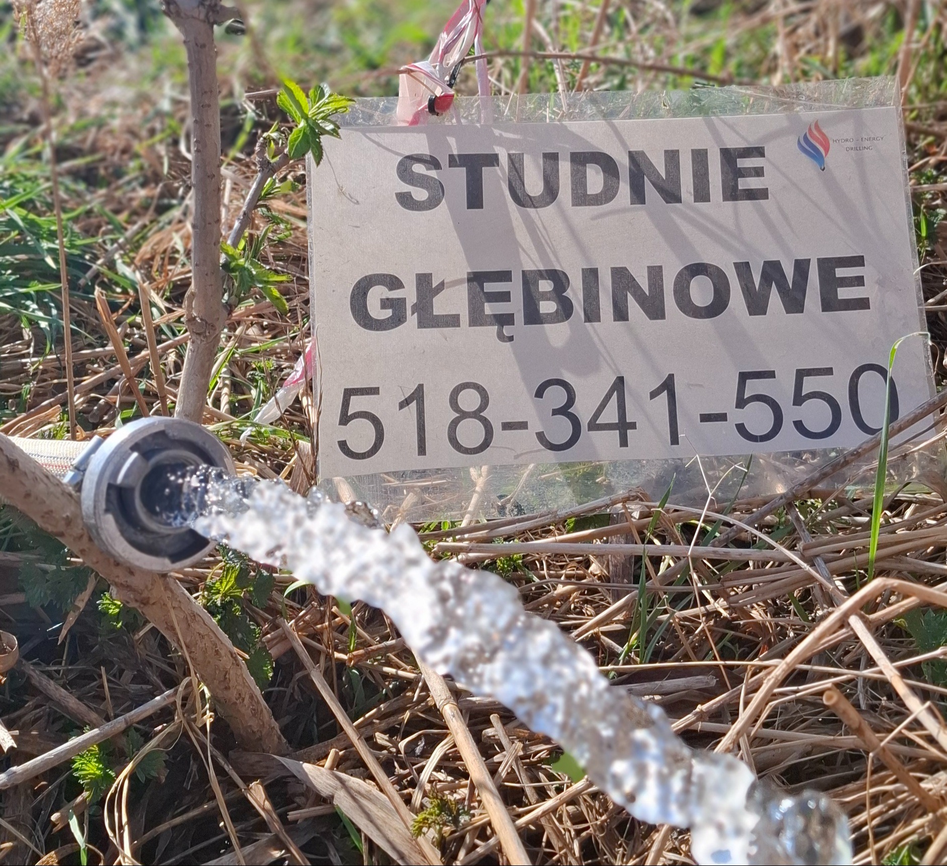 Woda wypływająca z węża na tle tablicy z numerem telefonu do firmy oferującej studnie głębinowe. Otoczenie to trawa i gałęzie.