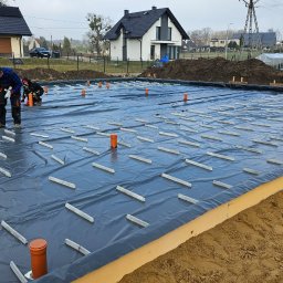 J.M.&S KONSTRUKCJE INŻYNIERSKIE I BUDOWLANE JACEK SZTENC - Fundamenty domu jednorodzinnego w trakcie budowy: izolacja przeciwwilgociowa, dystanse betonowe przed zbrojeniem płyty fundamentowej. Widoczni pracownicy w tle.
