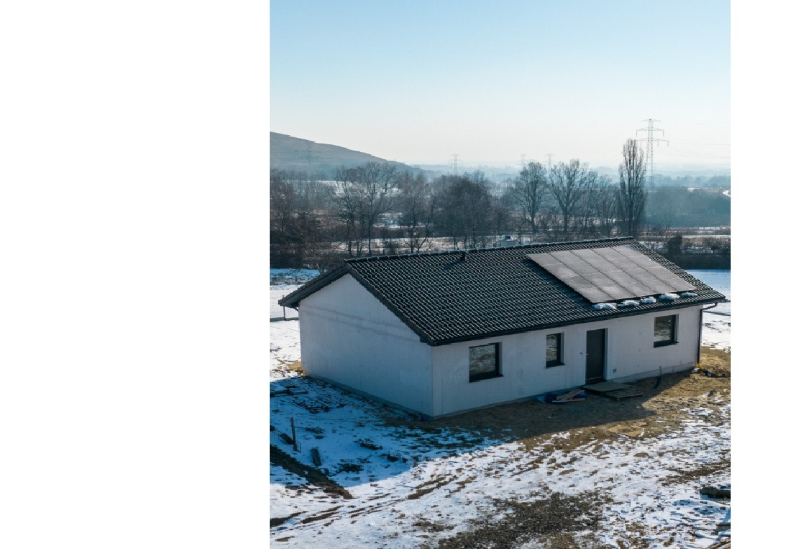 Dom prefabrykowany z panelami słonecznymi na dachu, w zimowej scenerii. Widok z góry ukazuje warstwowe ściany drewniane i konstrukcję KVH. W tle wzgórza i słupy wysokiego napięcia.