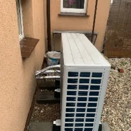 Zewnętrzna jednostka pompy ciepła zamontowana przy ścianie budynku z widocznymi rurami w izolacji, postawiona na betonowej podstawie i otoczona kamieniami.