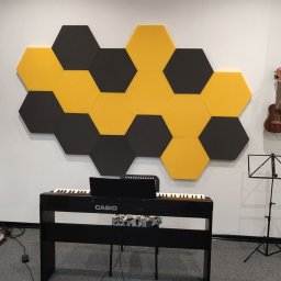 UNDERGROUND Damian Stroka - Pokój muzyczny z pianinem cyfrowym Casio, ukulele na ścianie, czarno-żółte panele akustyczne w kształcie heksagonów oraz statyw na nuty.