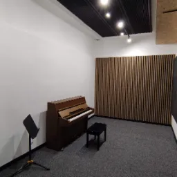 Wyciszona sala z pianinem, czarnym krzesłem i pulpitem na nuty. Panele akustyczne na ścianach i suficie, pionowe listwy dekoracyjne.