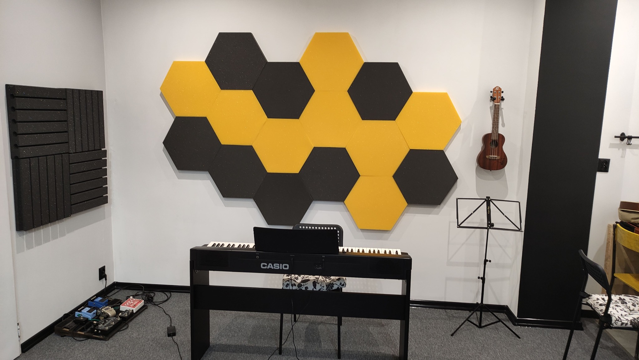 Pokój muzyczny z pianinem cyfrowym Casio, ukulele na ścianie, czarno-żółte panele akustyczne w kształcie heksagonów oraz statyw na nuty.