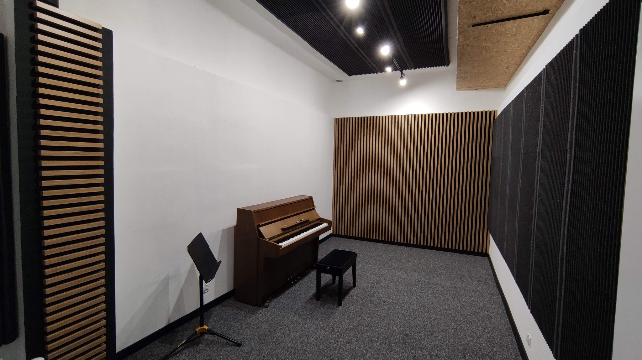 Wyciszona sala z pianinem, czarnym krzesłem i pulpitem na nuty. Panele akustyczne na ścianach i suficie, pionowe listwy dekoracyjne.