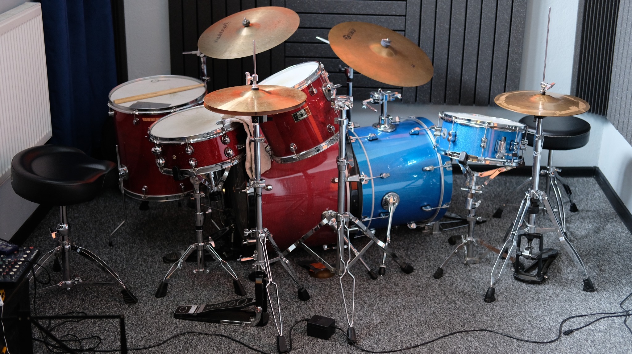 Zestaw perkusyjny w kolorach czerwonym i niebieskim, z talerzami, ustawiony w pomieszczeniu z akustycznymi panelami na ścianie.
