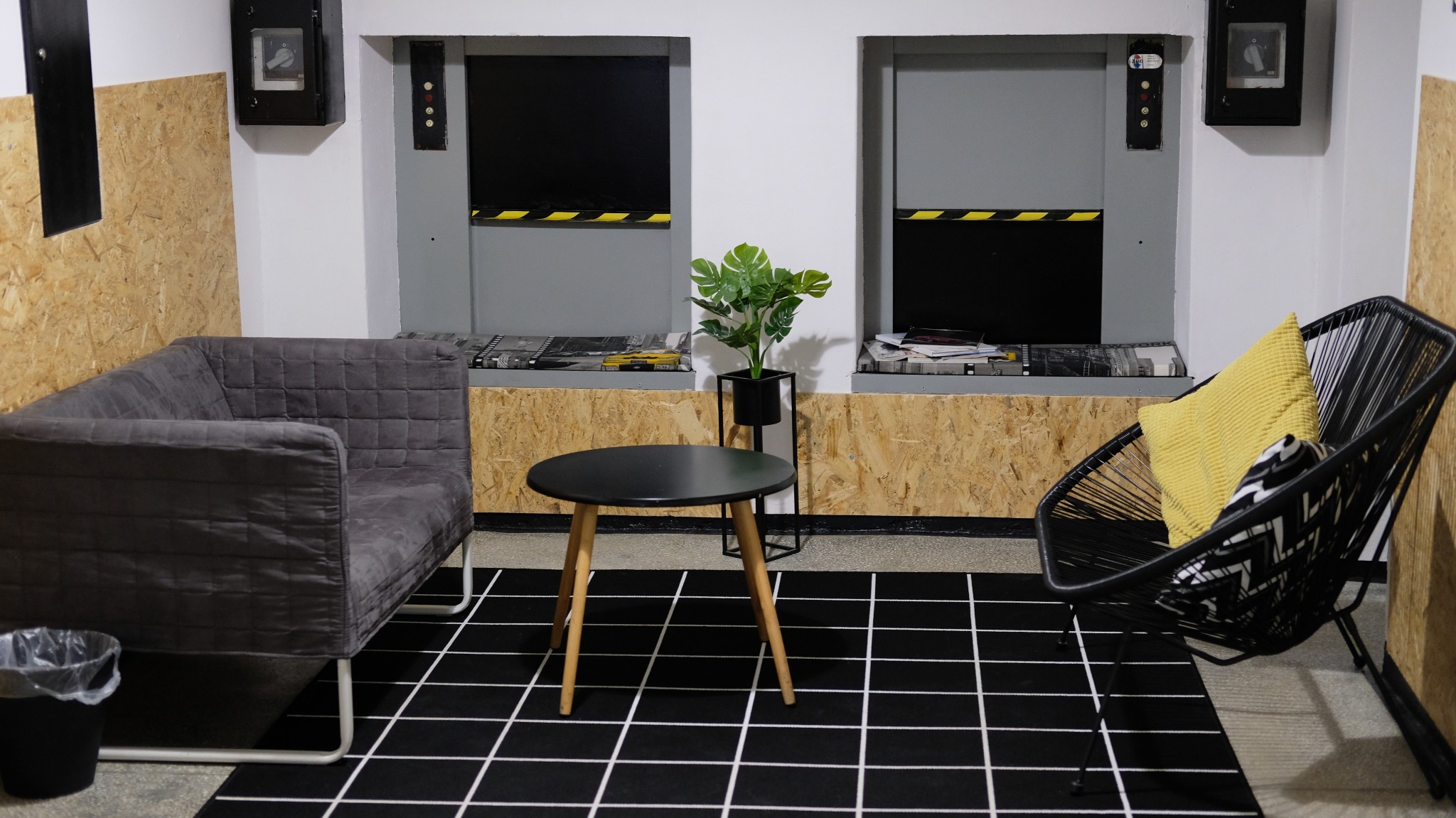 Wnętrze pokoju z szarą sofą, czarnym fotelem z żółtą poduszką, okrągłym stolikiem i rośliną doniczkową na tle ściany z płyt OSB i czarno-białą wykładziną w kratę.