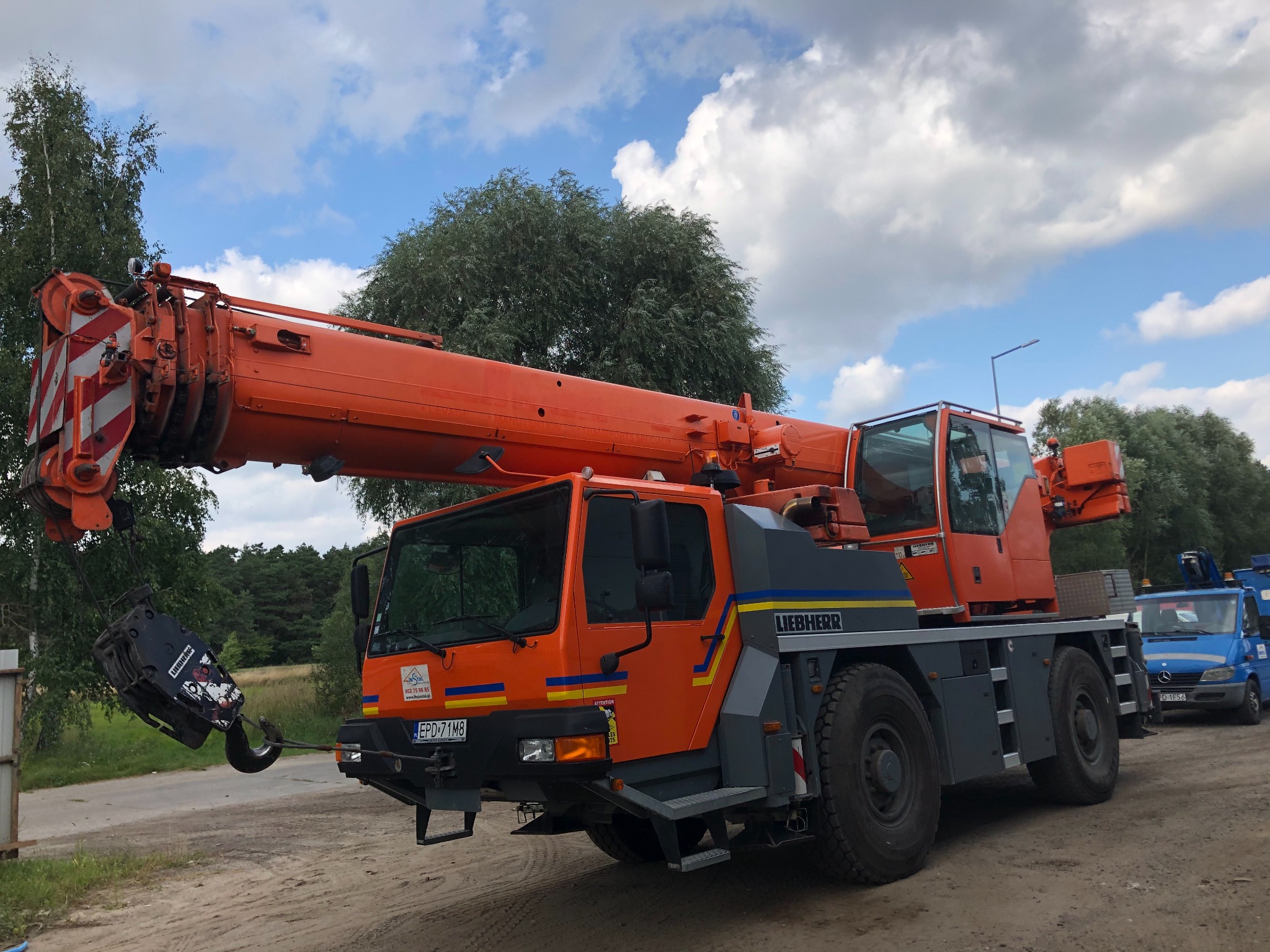 Pomarańczowy żuraw samochodowy Liebherr na placu budowy z hakiem i linami, na tle drzew i pochmurnego nieba.