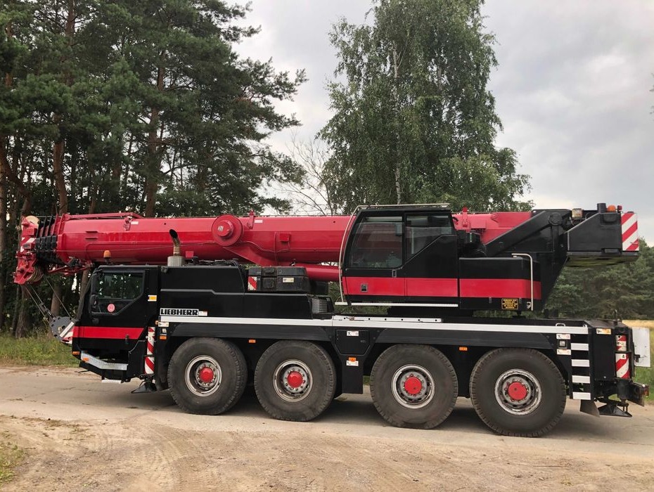 Czerwono-czarny dźwig samochodowy Liebherr na sześciu osiach, zaparkowany na żwirowej drodze, z lasem w tle.