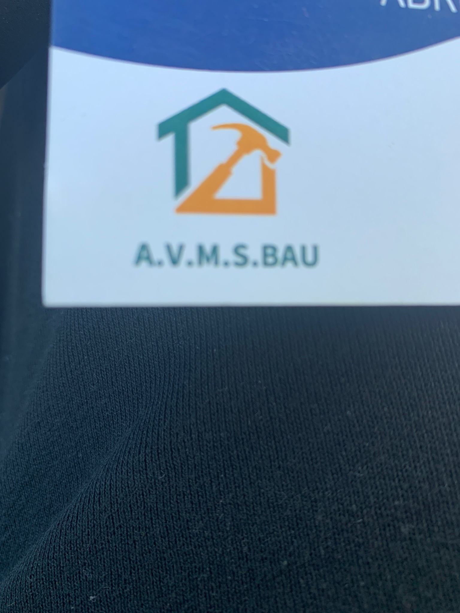 Wizytówka firmy A.V.M.S.BAU z logo przedstawiającym zarys domu i młotka w kolorach zielonym i pomarańczowym, na tle czarnej koszulki.