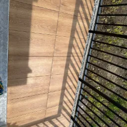 Widok z góry na balkon z drewnopodobną podłogą, czarną barierką i suszarką na pranie w tle, rzucający cień na płytki.