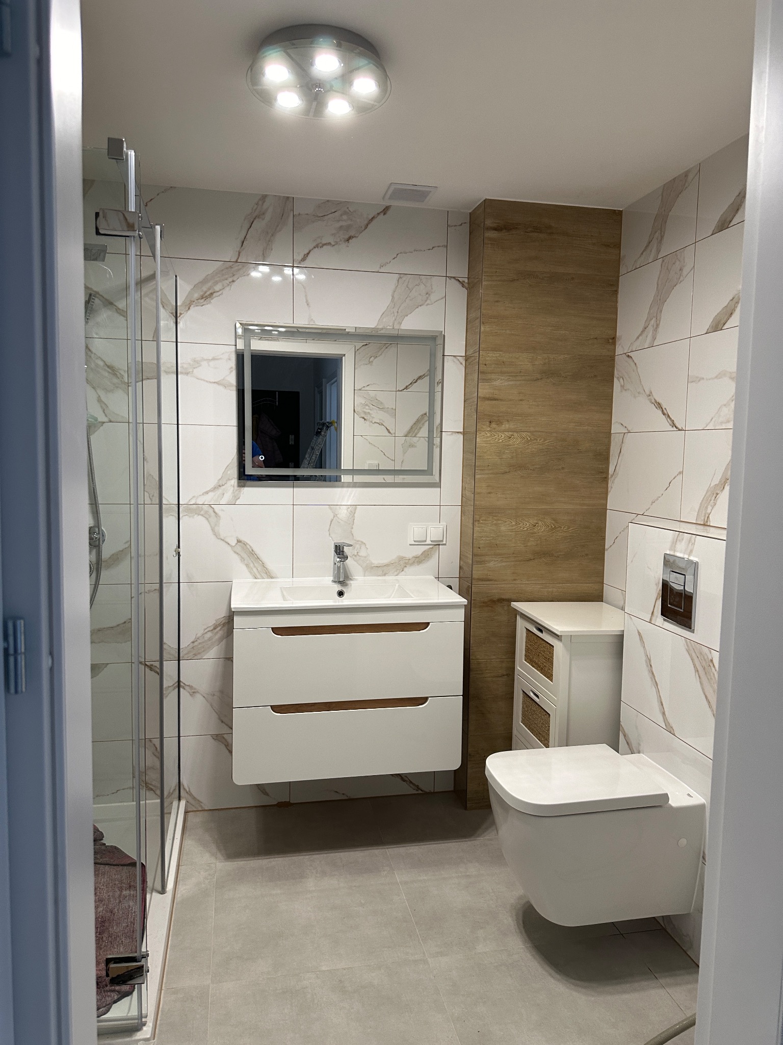 Nowoczesna łazienka z marmurowymi płytkami, wiszącą szafką z umywalką, toaletą podwieszaną i kabiną prysznicową z przezroczystego szkła.