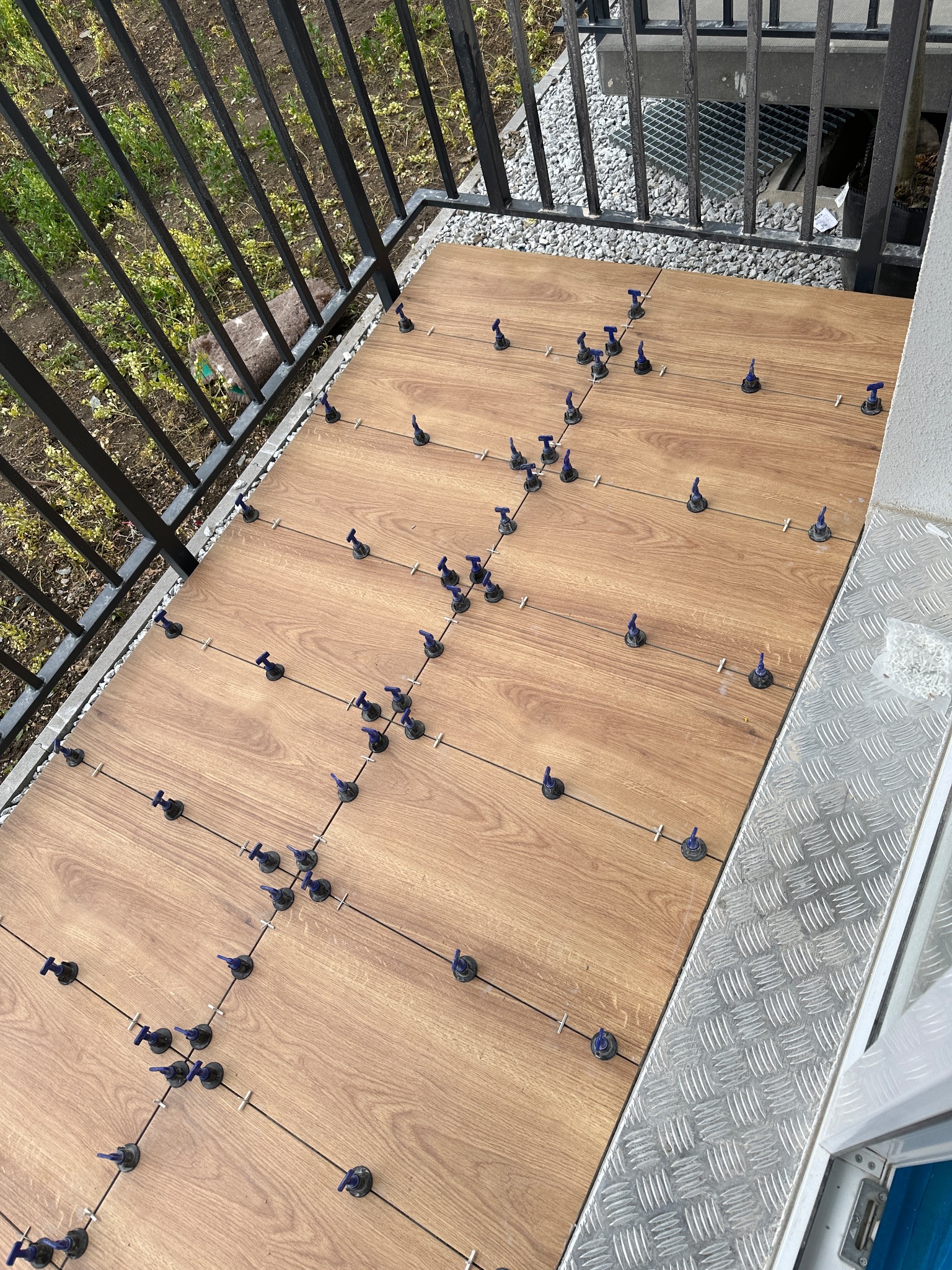 Układanie płytek imitujących drewno na balkonie z użyciem systemu poziomowania, widoczne krzyżyki dystansowe i zaciski, balustrada w tle.