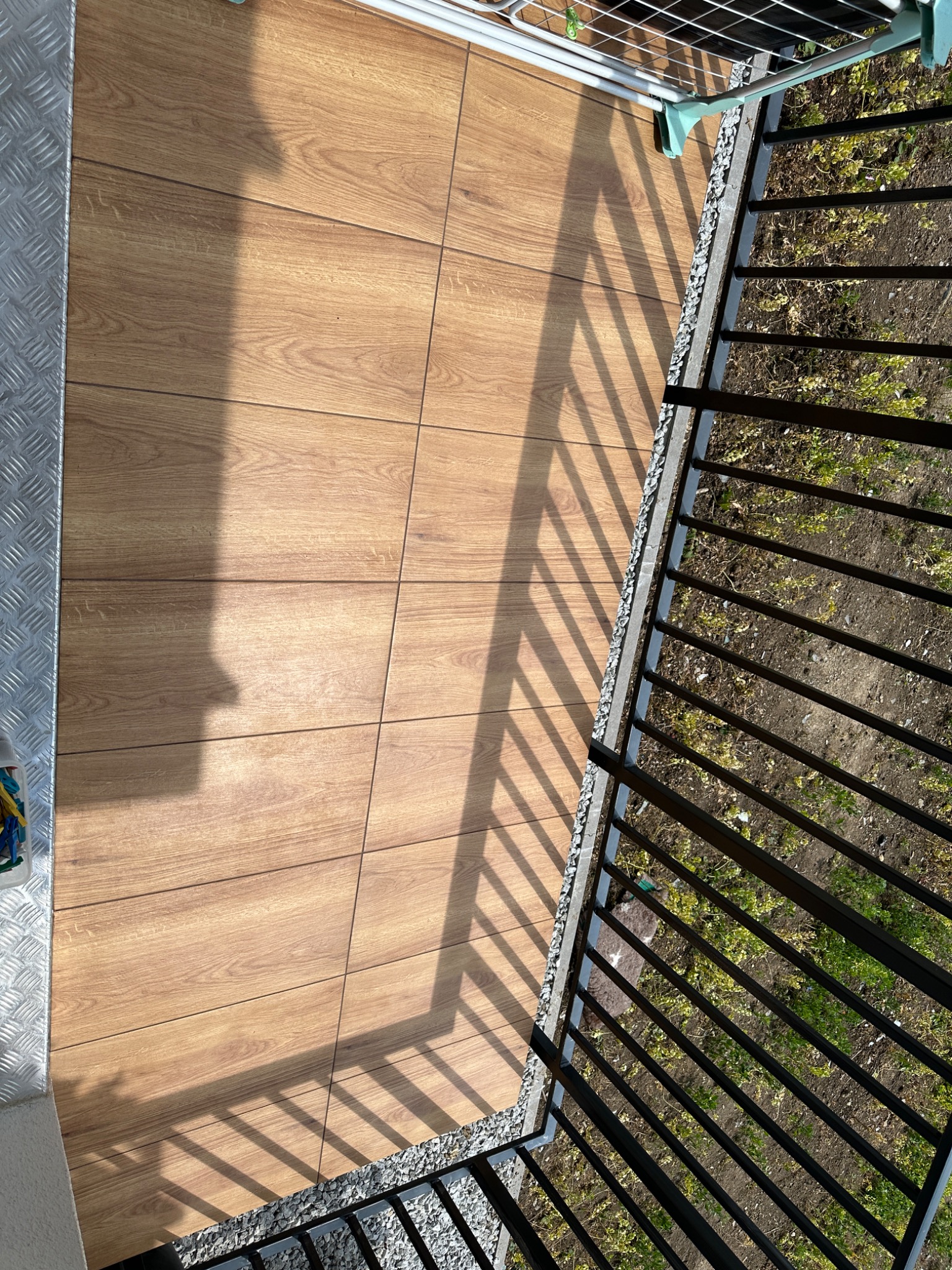 Widok z góry na balkon z drewnopodobną podłogą, czarną barierką i suszarką na pranie w tle, rzucający cień na płytki.