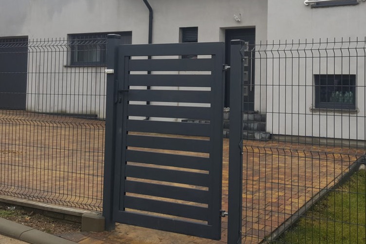 Otwarta, nowoczesna, grafitowa furtka z poziomymi panelami, stanowiąca część ogrodzenia panelowego, na tle domu z szarą elewacją i kostki brukowej.