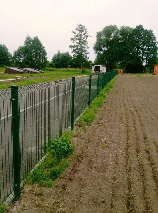 Długi odcinek zielonego ogrodzenia panelowego oddzielającego asfaltową drogę od zaoranego pola. W tle widoczny biały samochód dostawczy i drzewa.