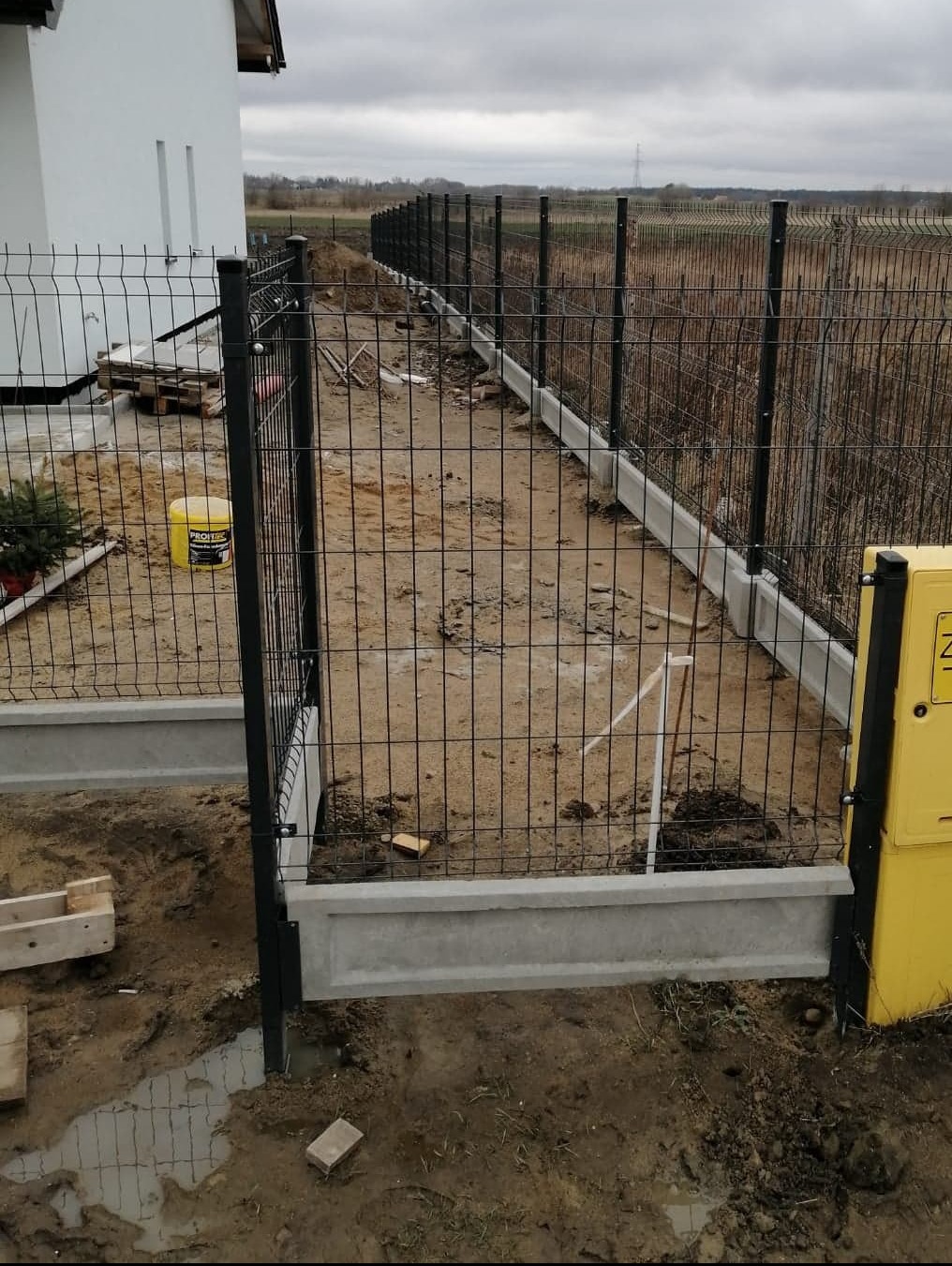 Montaż czarnego ogrodzenia panelowego na betonowej podmurówce, widoczne słupki i przęsła, teren po budowie, żółty pojemnik w tle.