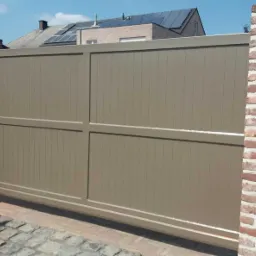 Automatyczna brama wjazdowa w kolorze beżowym, zamontowana między dwoma murowanymi filarami z czerwonej cegły, widok na dom z panelami słonecznymi na dachu.