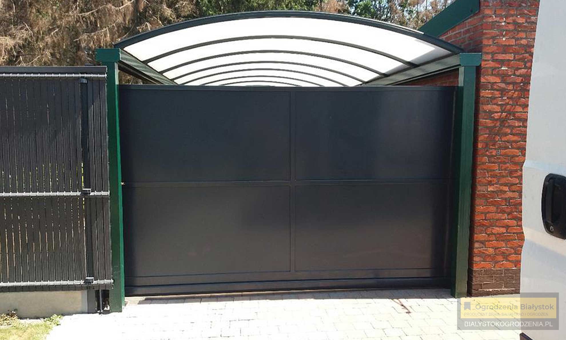 Nowoczesna, przesuwna brama wjazdowa w kolorze grafitowym, z zadaszeniem z poliwęglanu, widoczna z zewnątrz posesji, obok fragment ogrodzenia panelowego i mur z czerwonej cegły.