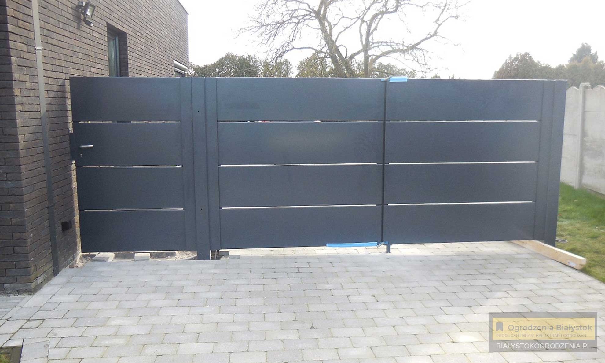 Nowoczesna, przesuwna brama wjazdowa w kolorze grafitowym z poziomymi, srebrnymi akcentami, zamontowana na kostce brukowej, z widocznym fragmentem ceglanego budynku w tle i zabezpieczeniami...