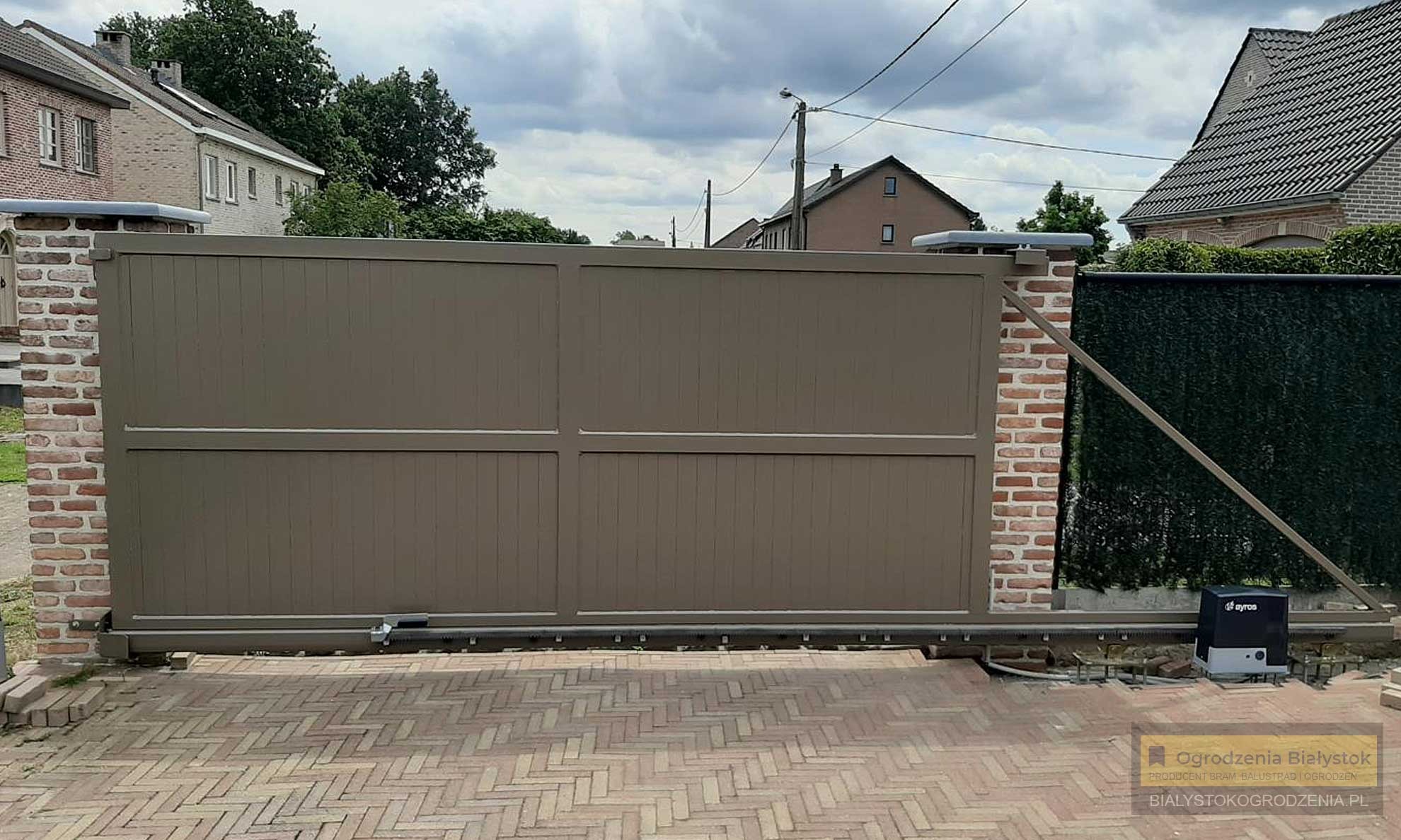 Przesuwna brama wjazdowa w kolorze taupe, zautomatyzowana, zamontowana na ceglanych słupkach, widoczny napęd i kostka brukowa na podjeździe.