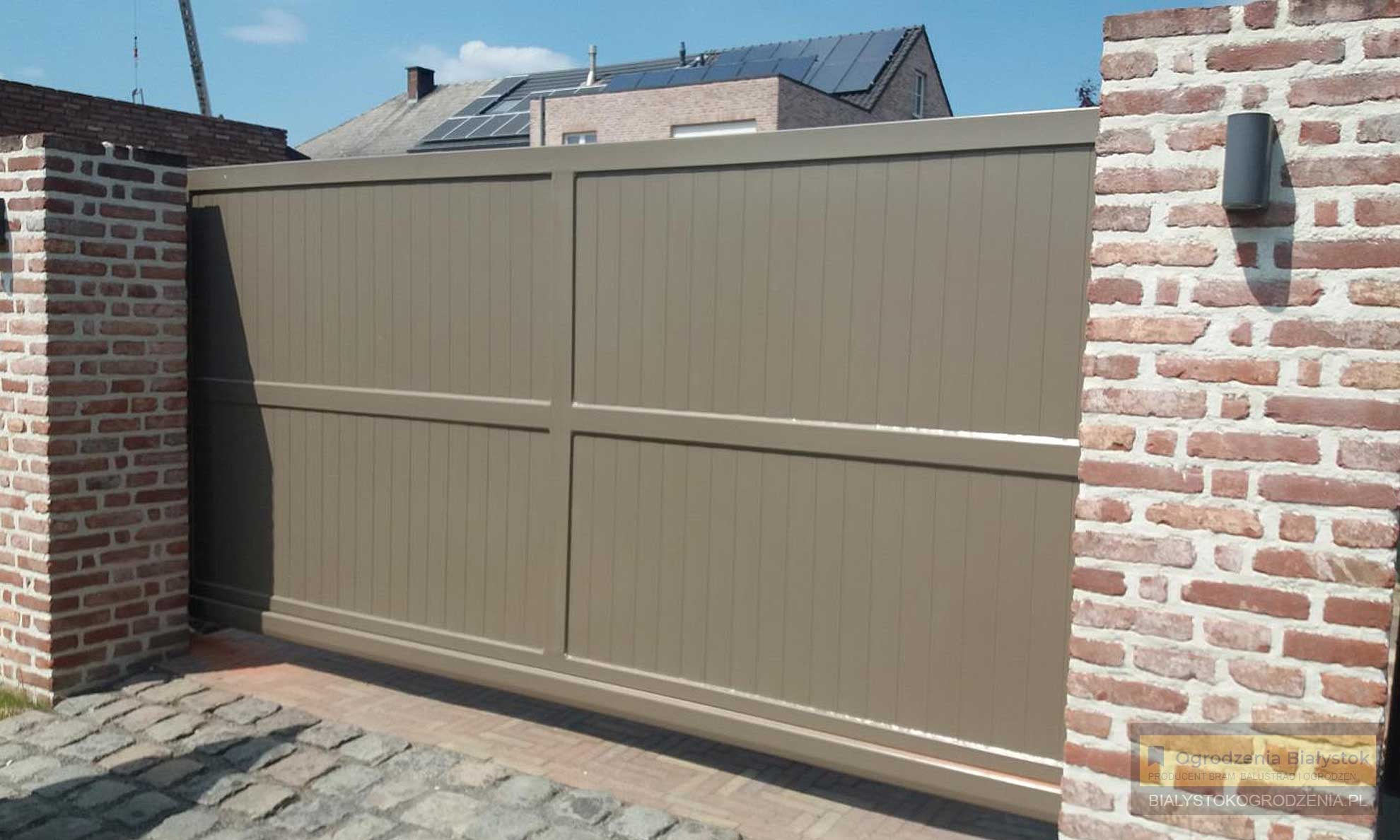 Automatyczna brama wjazdowa w kolorze beżowym, zamontowana między dwoma murowanymi filarami z czerwonej cegły, widok na dom z panelami słonecznymi na dachu.