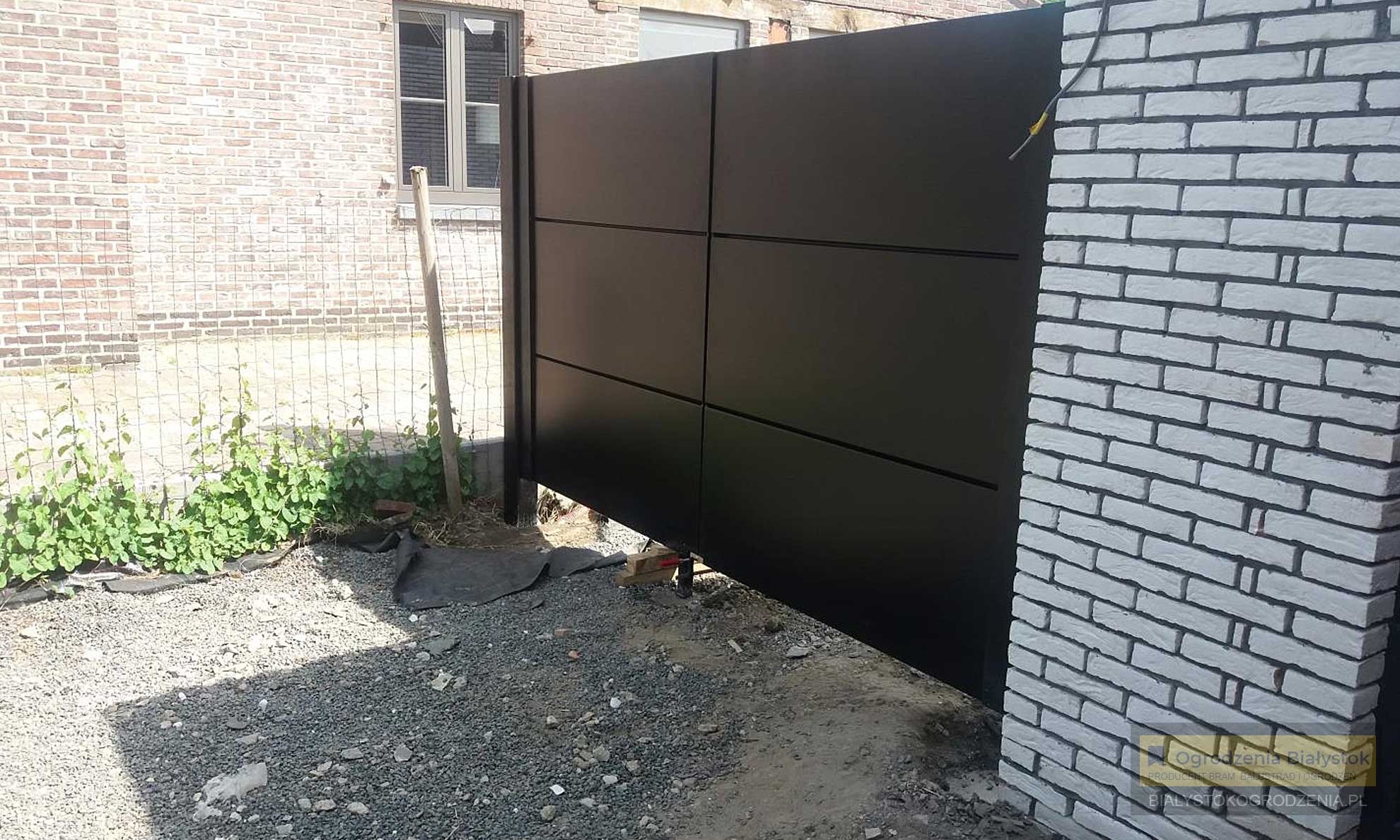 Nowoczesne, czarne ogrodzenie panelowe z widocznymi elementami montażowymi i częściowo wykończonym otoczeniem, kontrastujące z ceglanym murem.