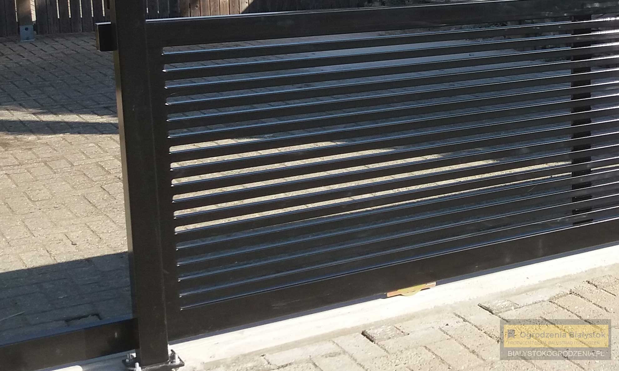 Nowoczesna, przesuwna brama wjazdowa z poziomych profili stalowych, w kolorze czarnym, widok z bliska na prowadnicę i betonową podstawę.