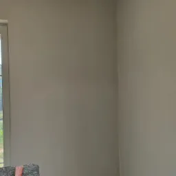 Świeżo pomalowane jasne ściany w pokoju, widok na zewnątrz przez okno z białymi ramami, fragment mebla w dolnej części kadru.