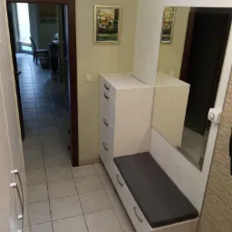 Przedpokój z zabudowaną szafką na buty z siedziskiem i lustrem, widok na korytarz i pokój z krzesłem przez otwarte drzwi.