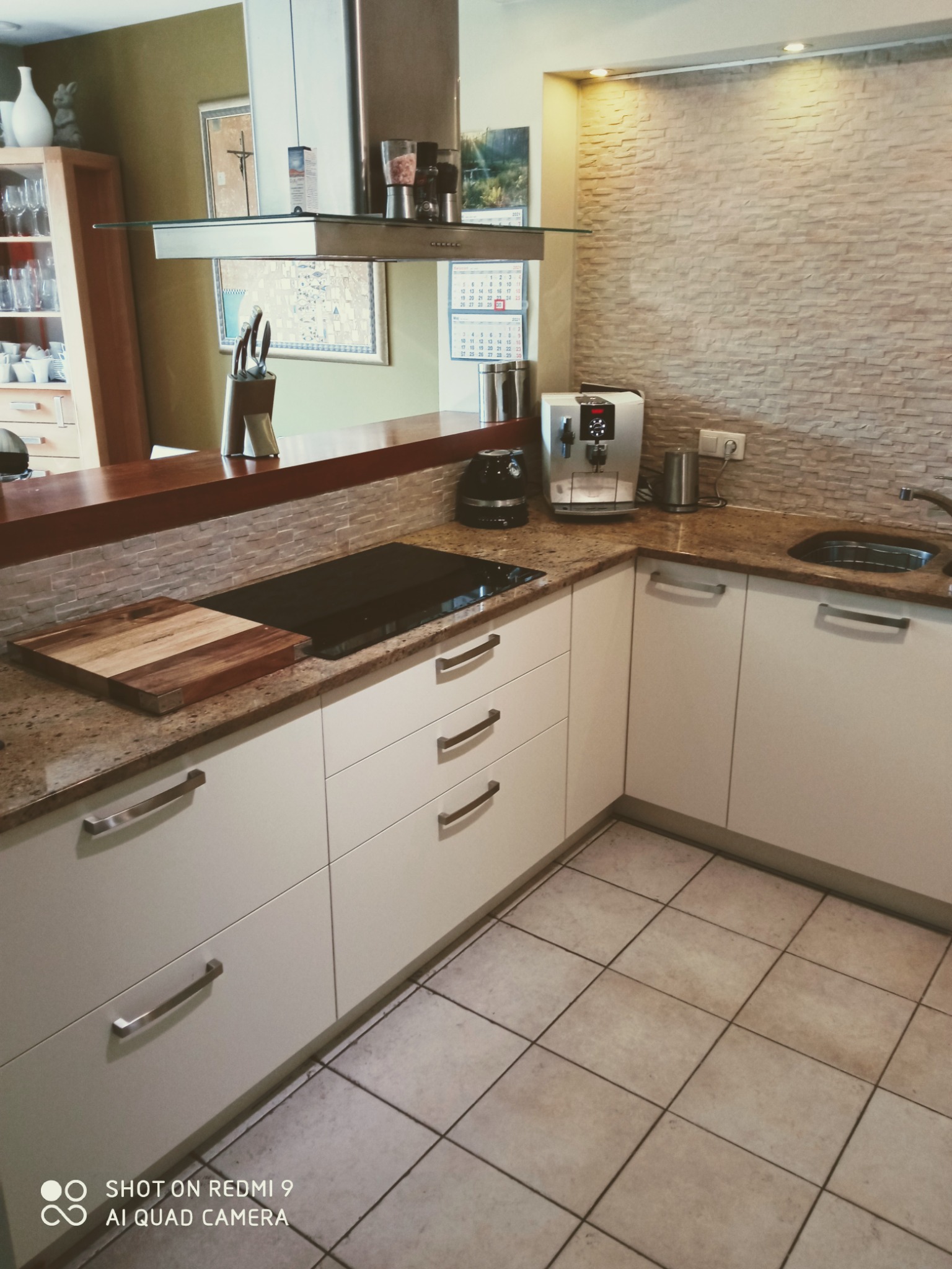 Jasna kuchnia z białymi szafkami, blatem z jasnego kamienia i kamienną ścianą, wyposażona w płytę grzewczą, ekspres do kawy i drewnianą deskę do krojenia.