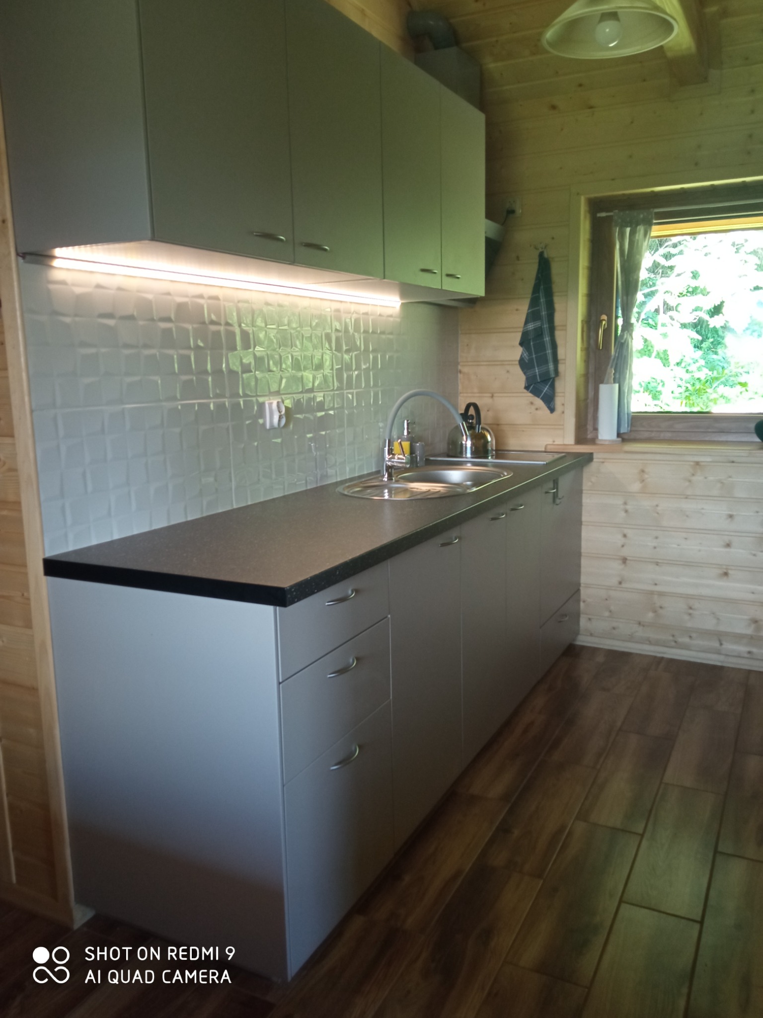Kuchnia w domku drewnianym: szare szafki kuchenne z ciemnym blatem, biała mozaika na ścianie, oświetlenie LED pod szafkami, widok z okna na zieleń.
