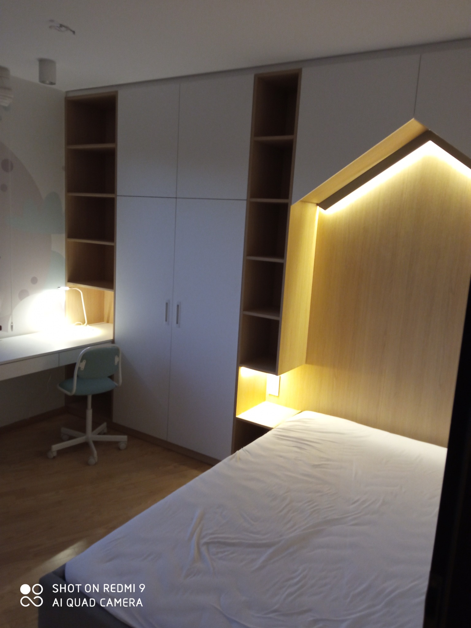 Pokój z łóżkiem z oświetleniem LED w zagłówku, biurkiem z krzesłem i szafą wnękową z półkami.