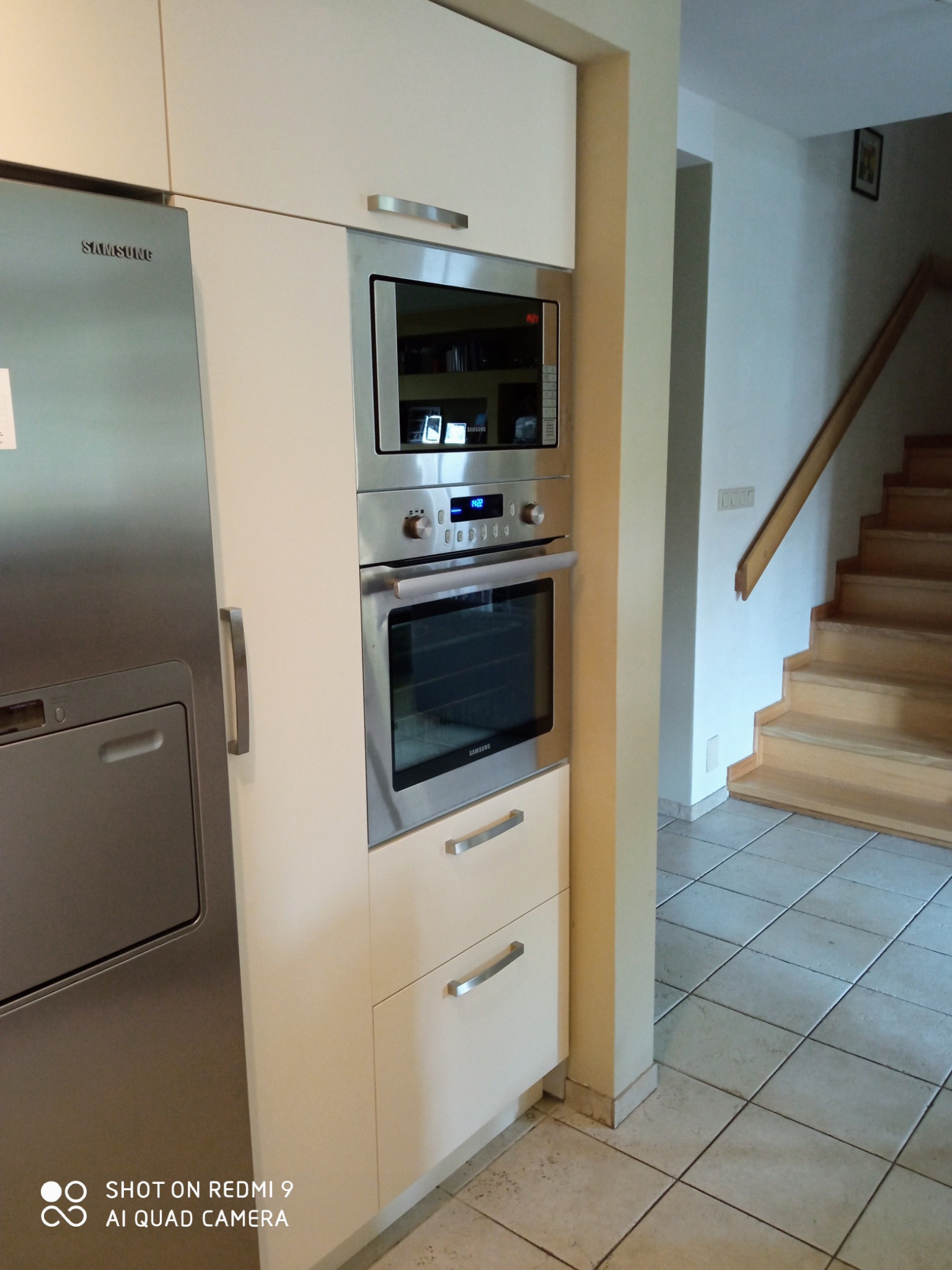 Biała zabudowa kuchenna z piekarnikiem, mikrofalówką i szufladami, z widoczną lodówką Samsung po lewej stronie i fragmentem drewnianych schodów w tle.