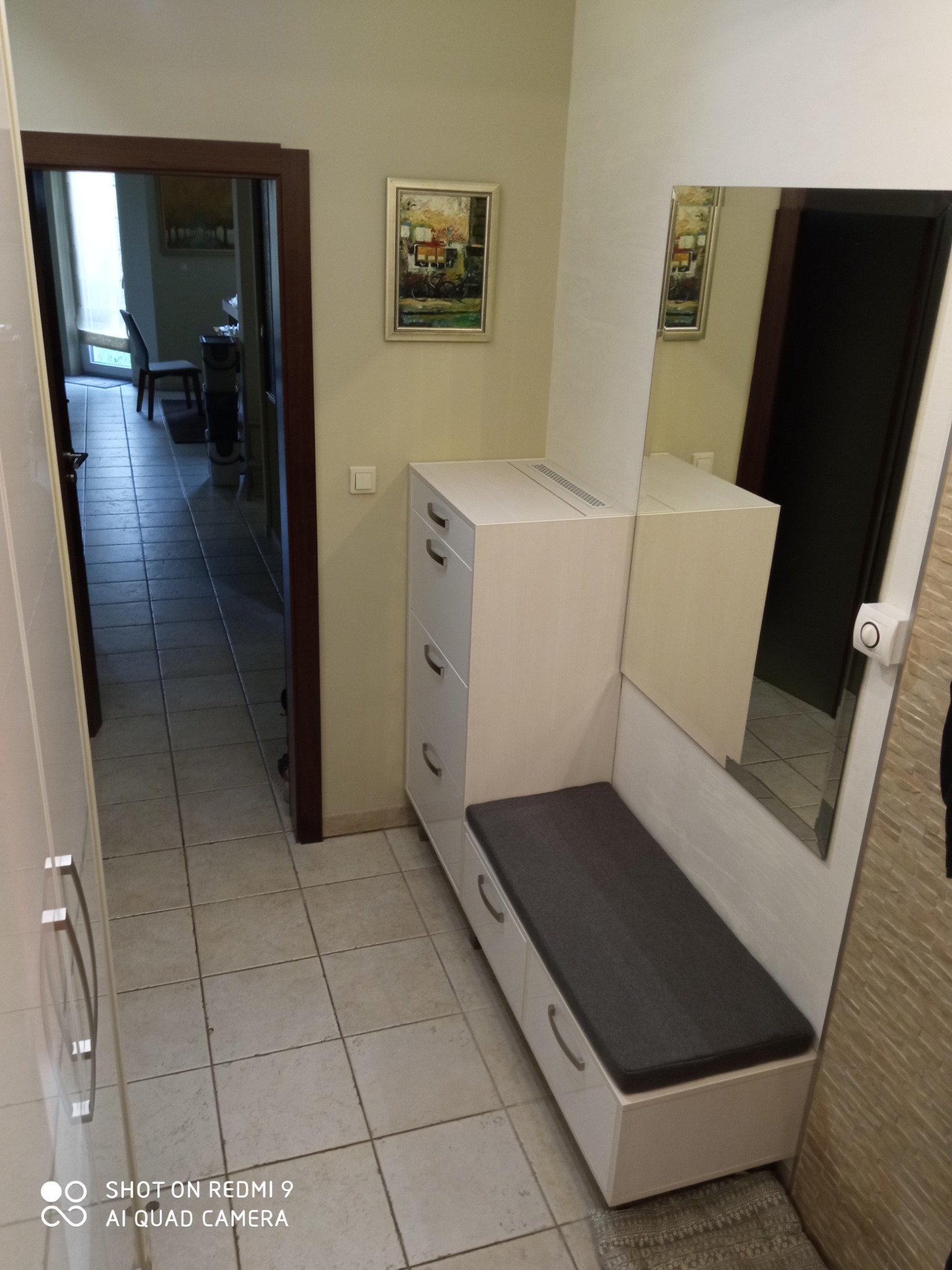 Przedpokój z zabudowaną szafką na buty z siedziskiem i lustrem, widok na korytarz i pokój z krzesłem przez otwarte drzwi.