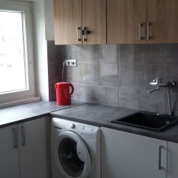 Pracownia - Szare płytki na ścianie nad blatem kuchennym z czarnym zlewozmywakiem, obok czerwony czajnik elektryczny i pralka automatyczna, nad blatem wiszące szafki z jasnego drewna.