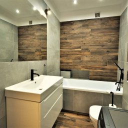 Pracownia - Nowoczesna łazienka z drewnopodobnymi płytkami na ścianie, szarą armaturą i białymi meblami łazienkowymi, widok na umywalkę, wannę i toaletę.