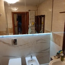 Pracownia - Wykończona łazienka z toaletą, bidetem i umywalką na tle marmurowych płytek, oświetlona listwą LED pod lustrem.