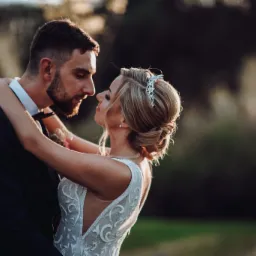 Para młoda w objęciach, panna młoda w sukni z koronkowymi zdobieniami, pan młody w ciemnym garniturze z muszką, delikatne oświetlenie zza drzew.