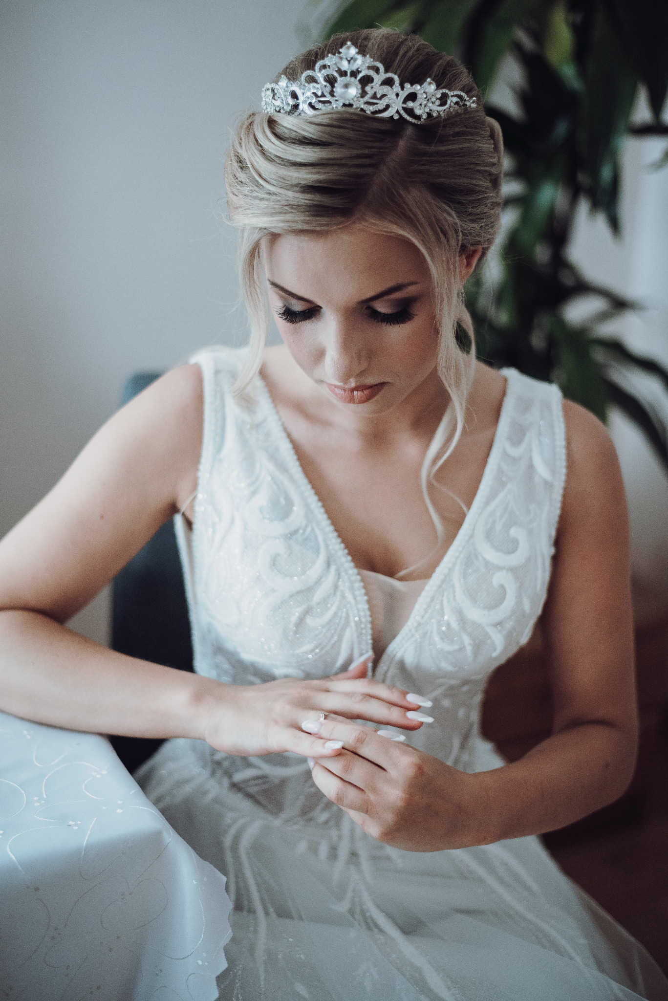 Panna młoda w białej sukni z tiarą, spogląda na dłonie z pierścionkiem zaręczynowym, delikatne światło.