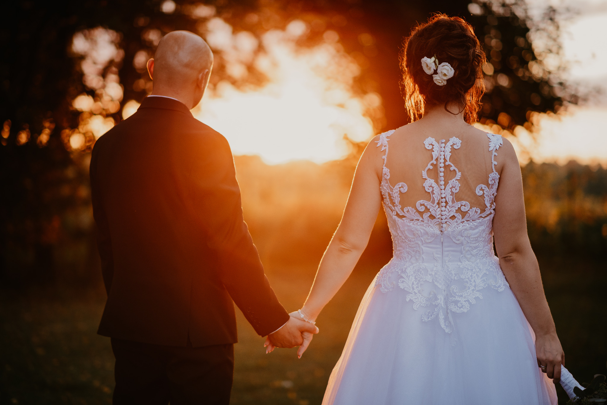 Para młoda trzymająca się za ręce, widok z tyłu, suknia z koronkowymi zdobieniami na plecach, zachód słońca w tle.