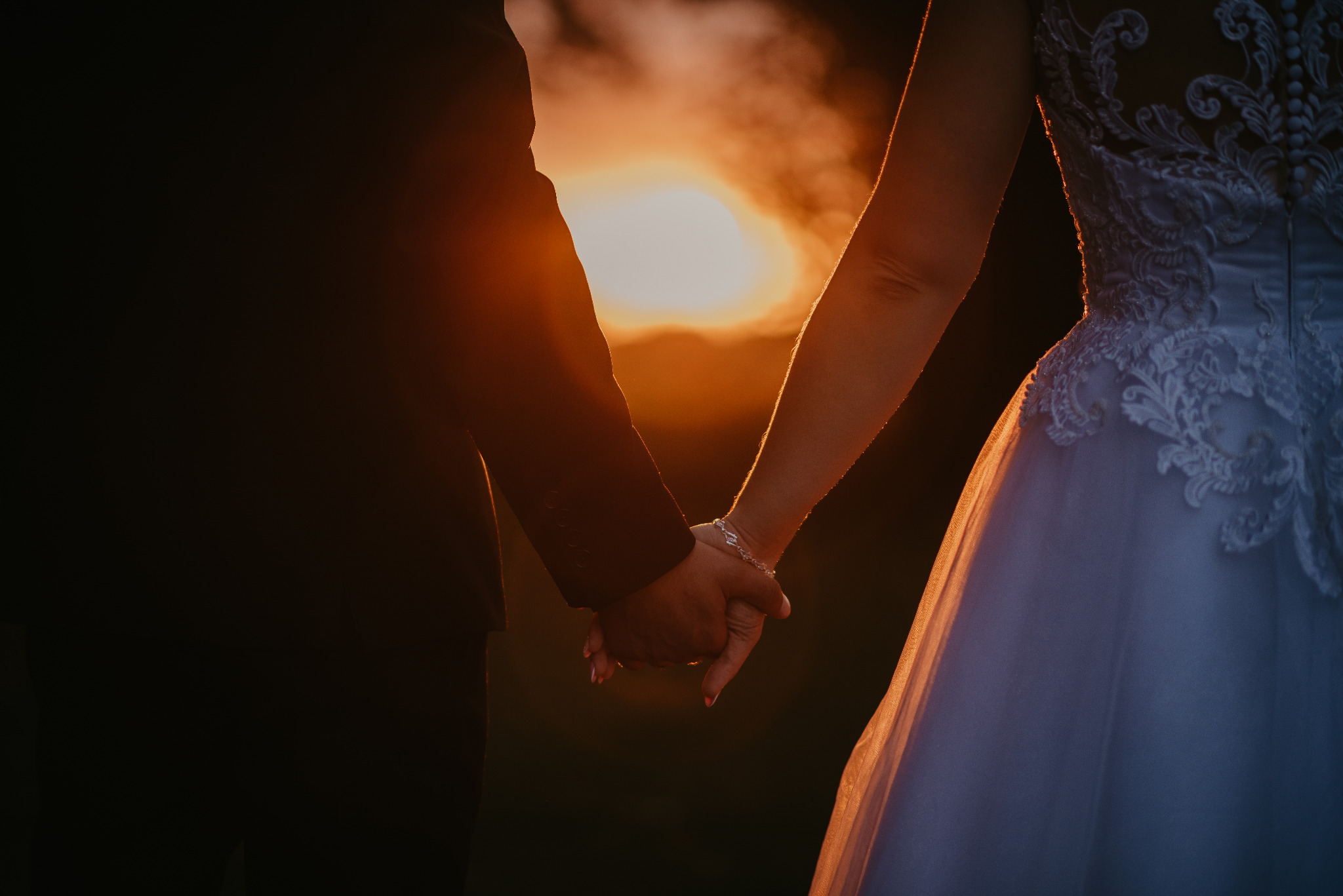 Para trzymająca się za ręce na tle zachodzącego słońca w Gdyni; widoczny fragment sukni ślubnej z koronkowym wykończeniem i bransoletka na nadgarstku panny młodej.